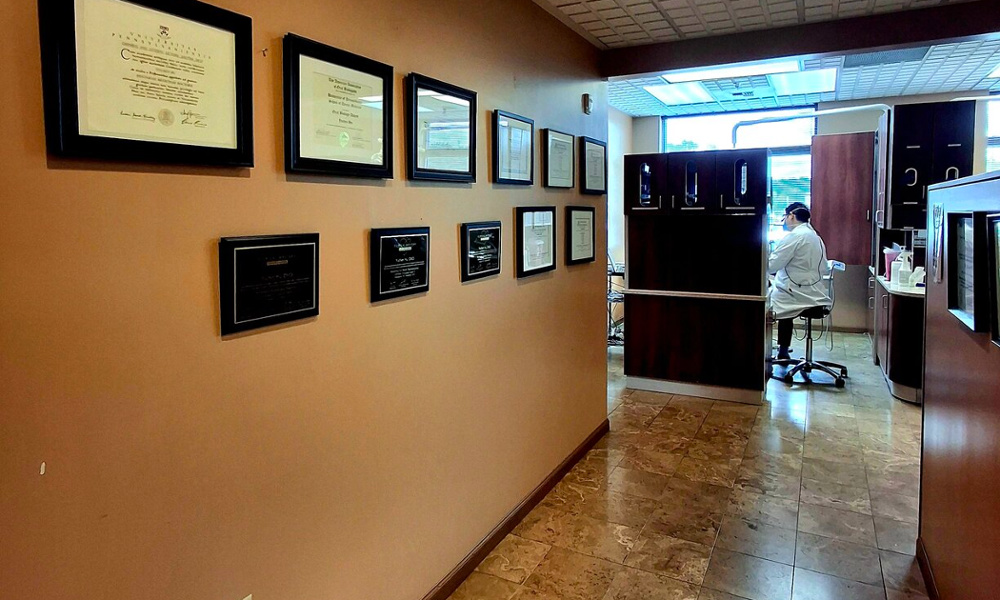 Dental office hallway with framed credentials and diplomas on warm-toned walls, staff member working at desk in background